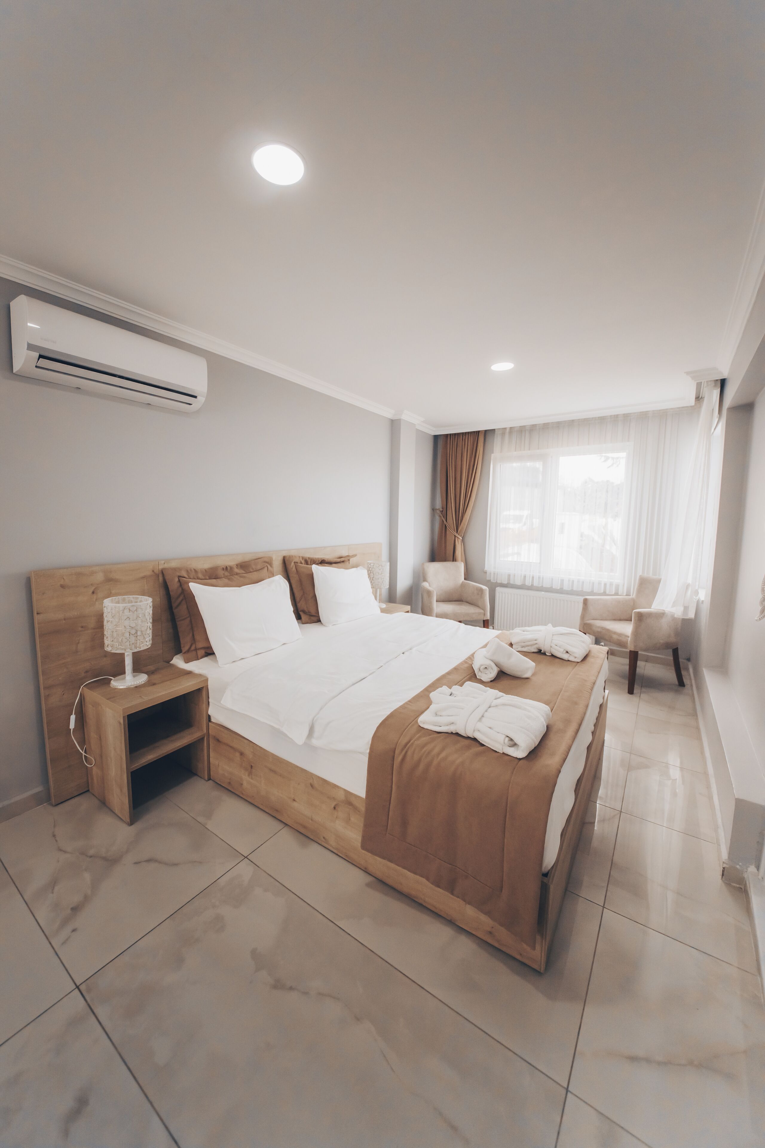 A modern hotel room with a wooden bed, white linens, a bedside table with a lamp, and a window with curtains.