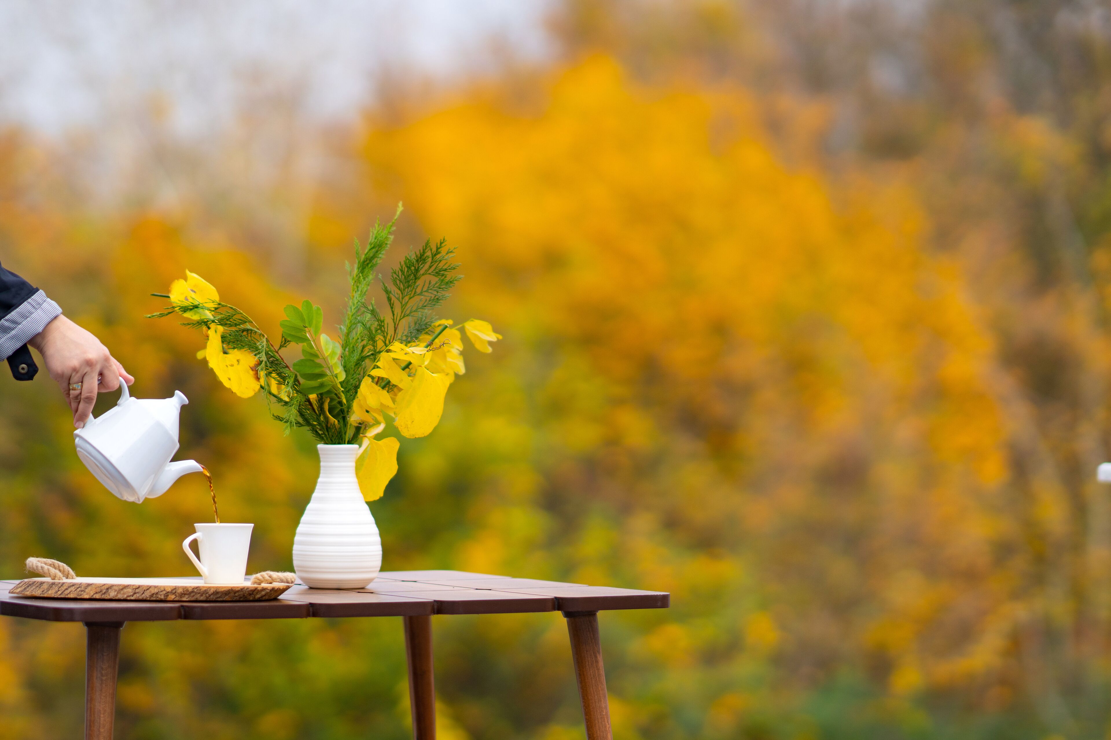 En hvid tepotte hælder i en hvid kop på et træbord med en hvid vase med gule blomster.