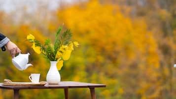 Una tetera blanca vertiendo en una taza blanca sobre una mesa de madera con un florero blanco que contiene flores amarillas.