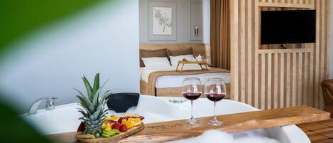 A hotel room with a bathtub, a fruit platter, two glasses of red wine, and a bed with a wooden headboard.