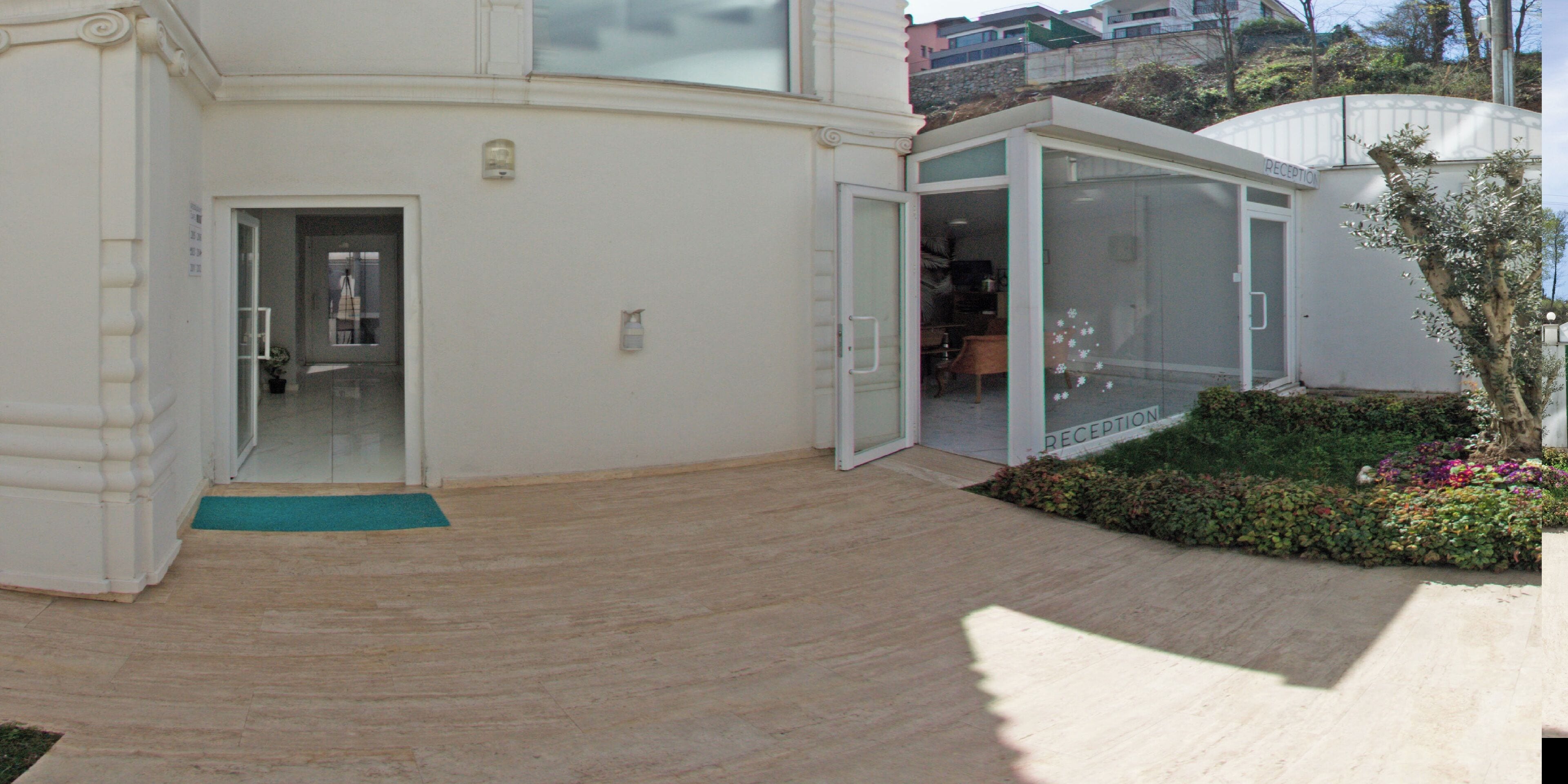 Zona de recepción con puerta de vidrio, mostrador de recepción y un pequeño jardín con plantas y flores.