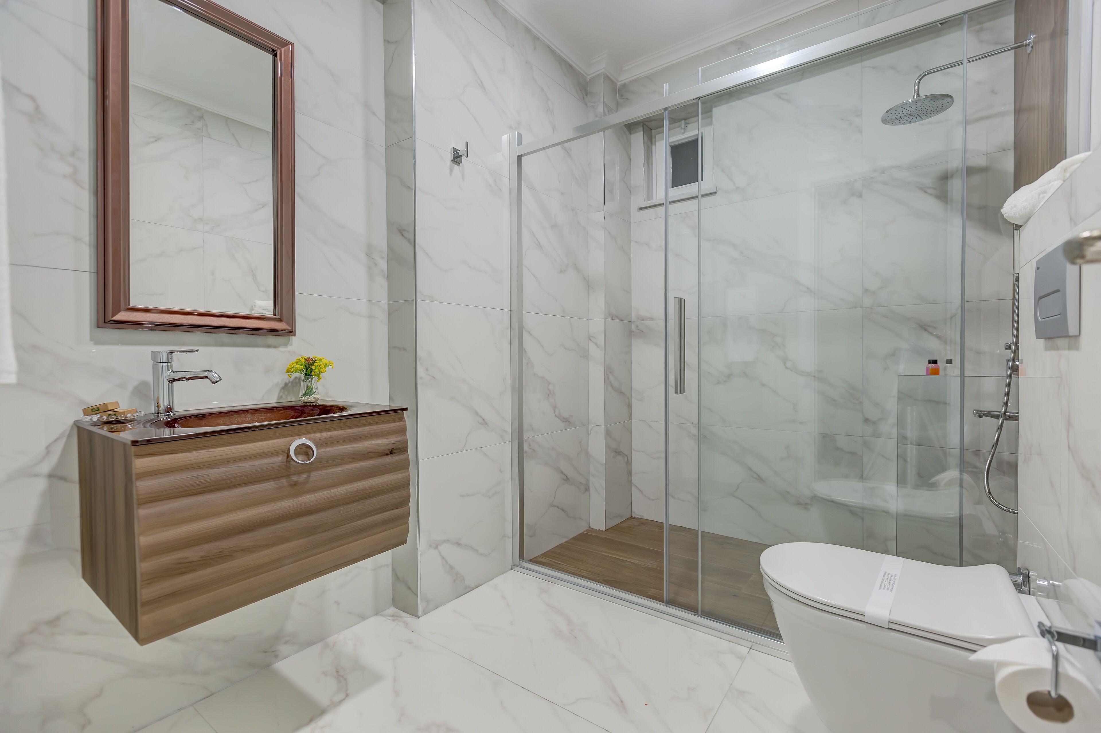 A modern bathroom with a marble floor, a wooden vanity, a glass shower enclosure, and a toilet.