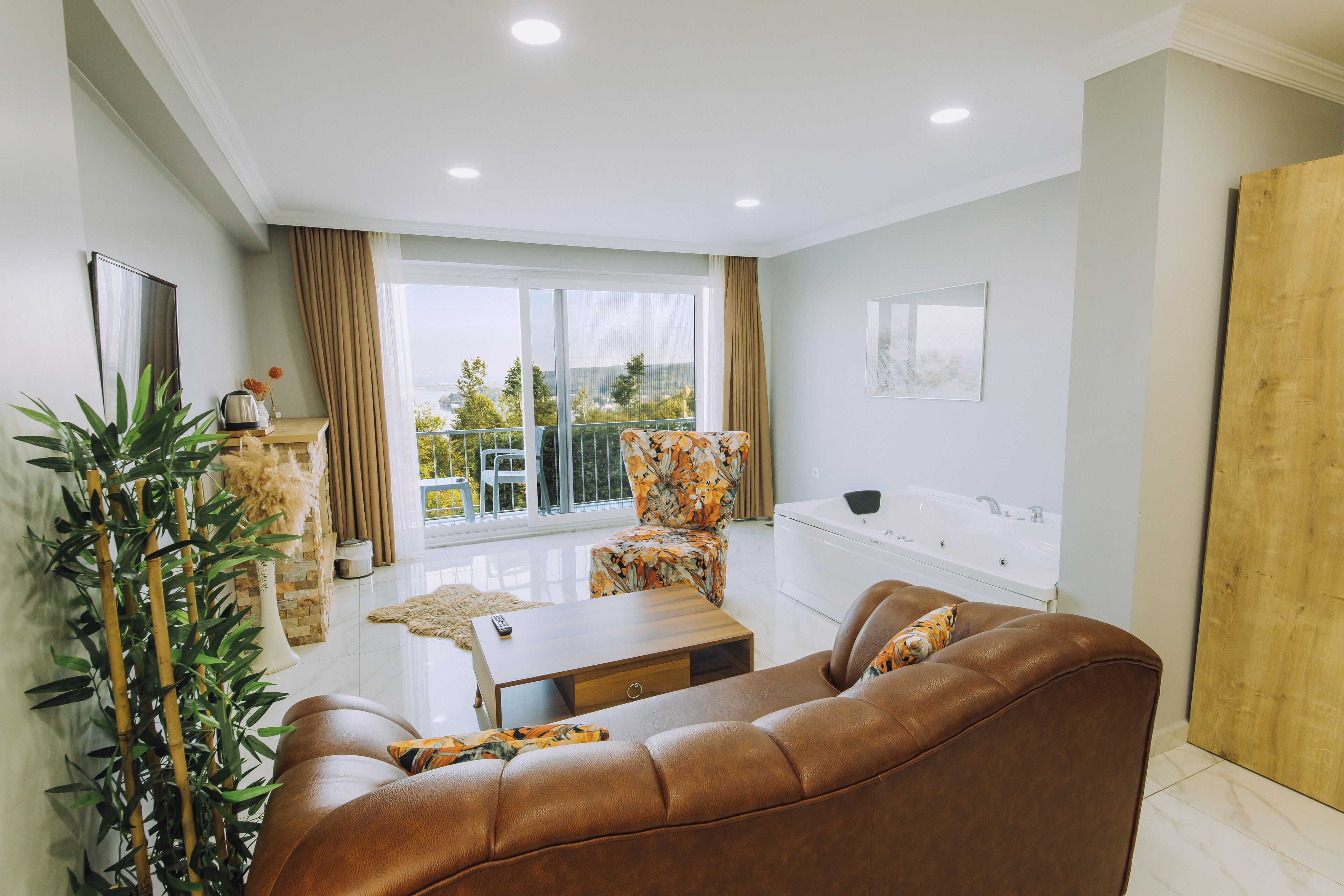 A modern living room with a brown leather sofa, a wooden coffee table, and a floral armchair. There is a large window with a view of greenery and a balcony.