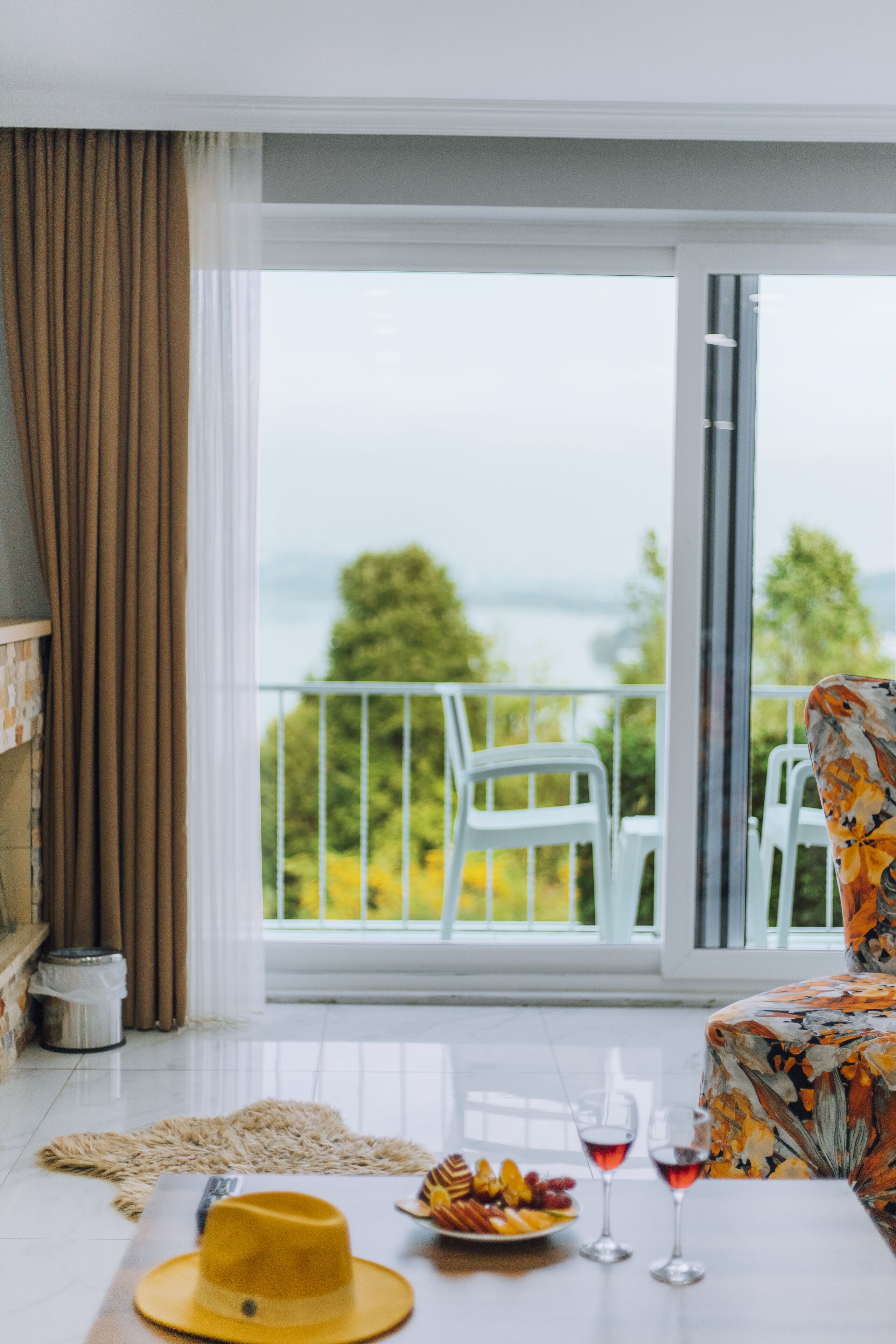 A room with a balcony, a dining table with a hat and fruit, and two wine glasses.