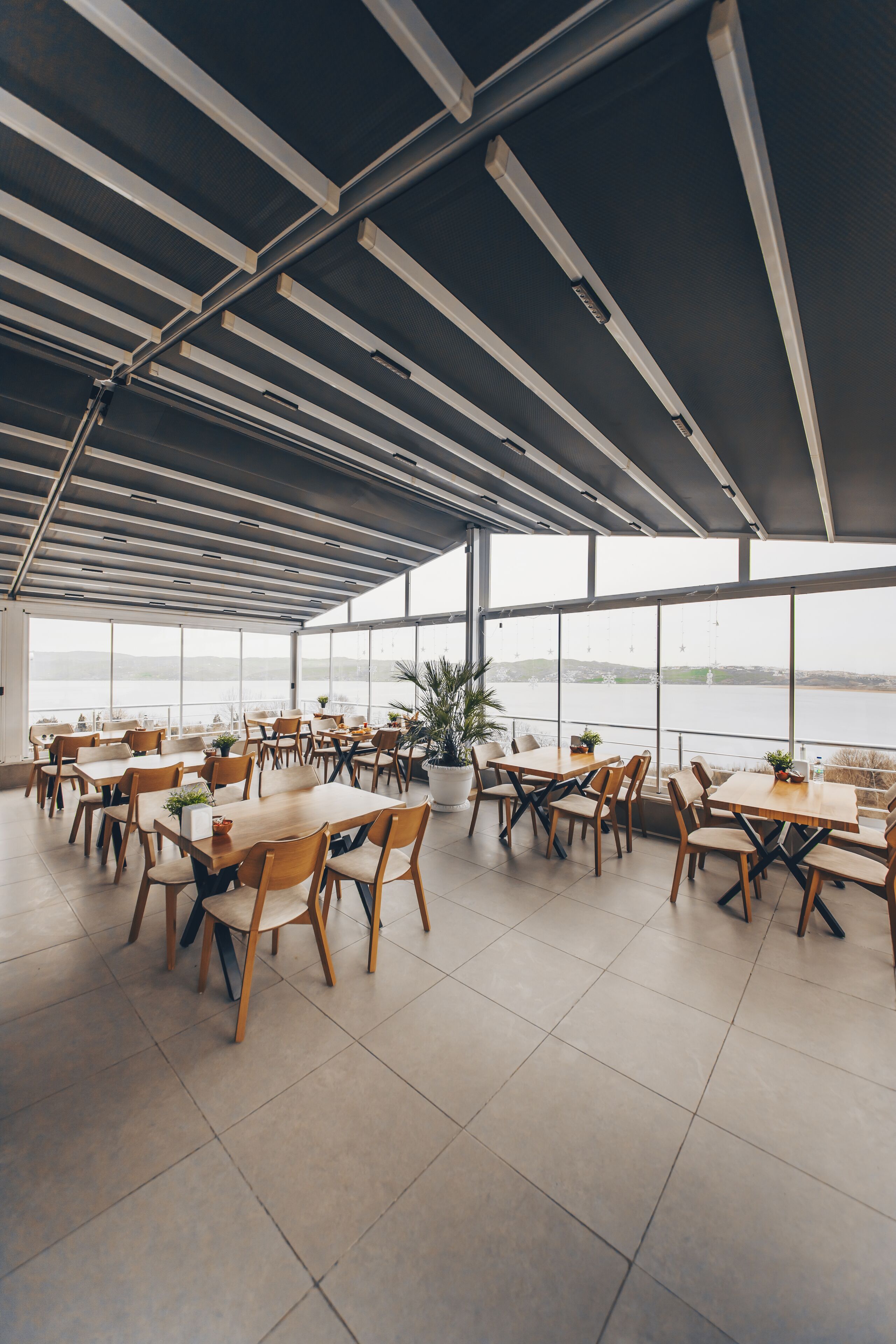 A spacious indoor dining area with large windows overlooking a body of water, featuring wooden tables and chairs.