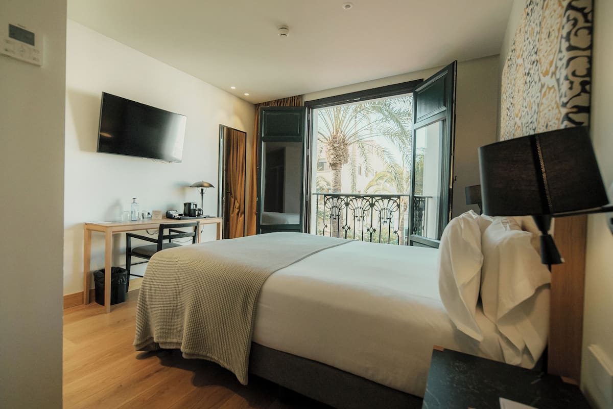 A hotel room with a bed, a desk, a TV, and a window with a view of palm trees.