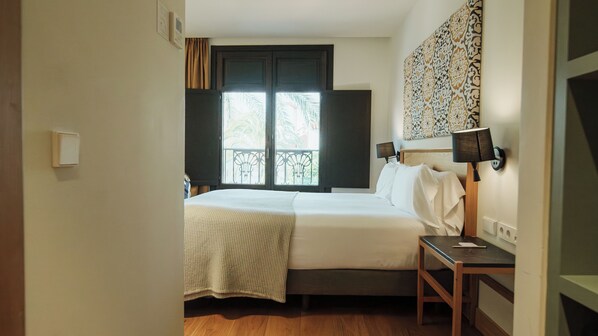Un dormitorio con cama, mesita de noche, lámpara y ventana con vistas a palmeras.