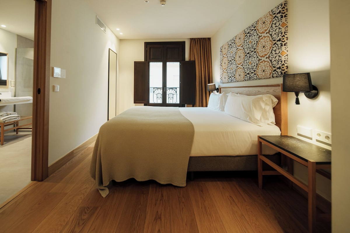 A hotel room with a large bed, a bedside table, a mirror, a window with curtains, and a wall-mounted lamp.