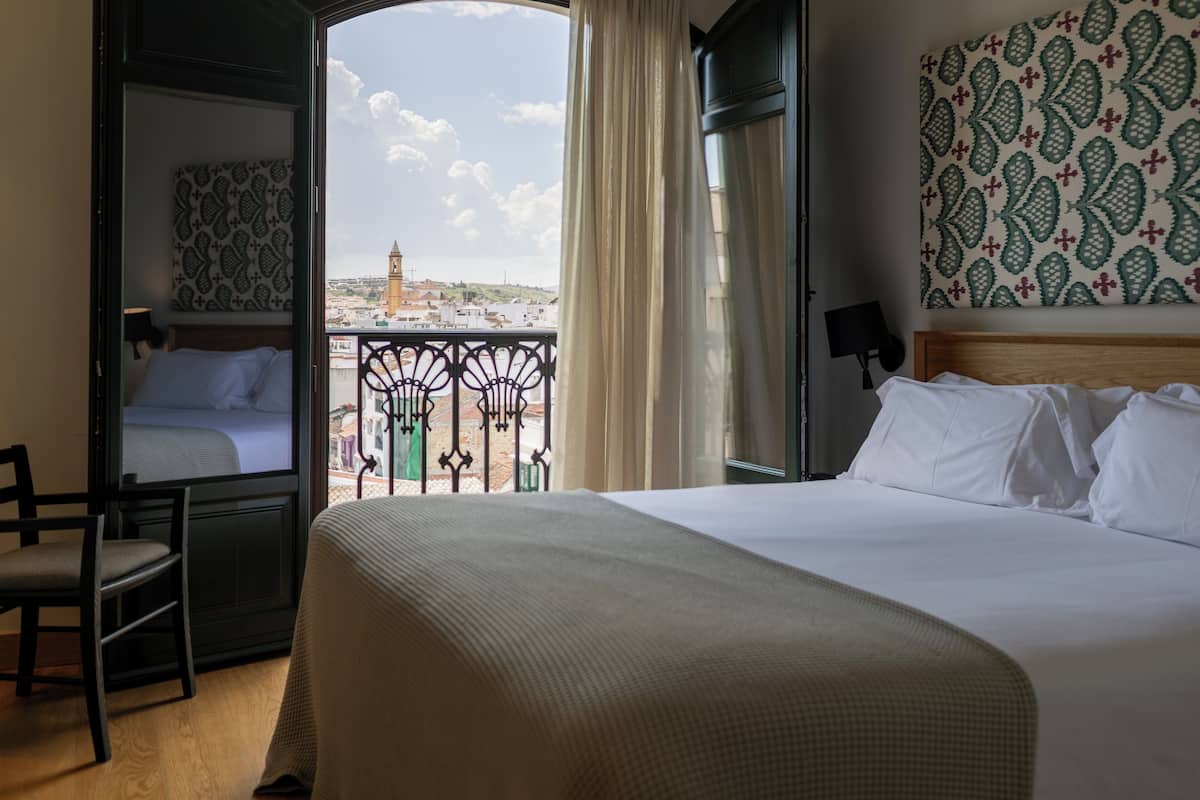 A hotel room with a large bed, a chair, a view of a cityscape, and a balcony with a railing.