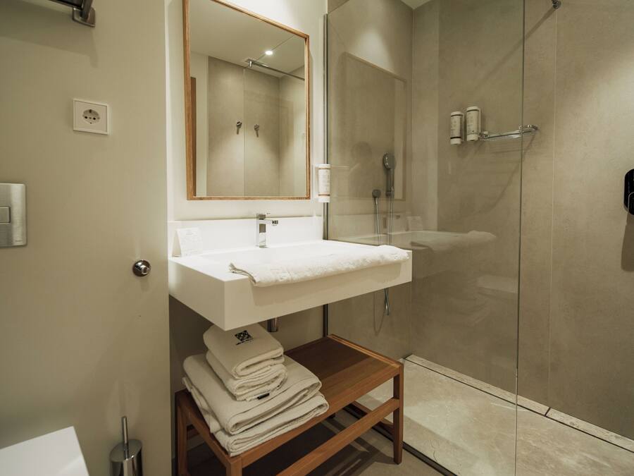 Un baño moderno con ducha acristalada, lavamanos blanco y estante de madera con toallas.