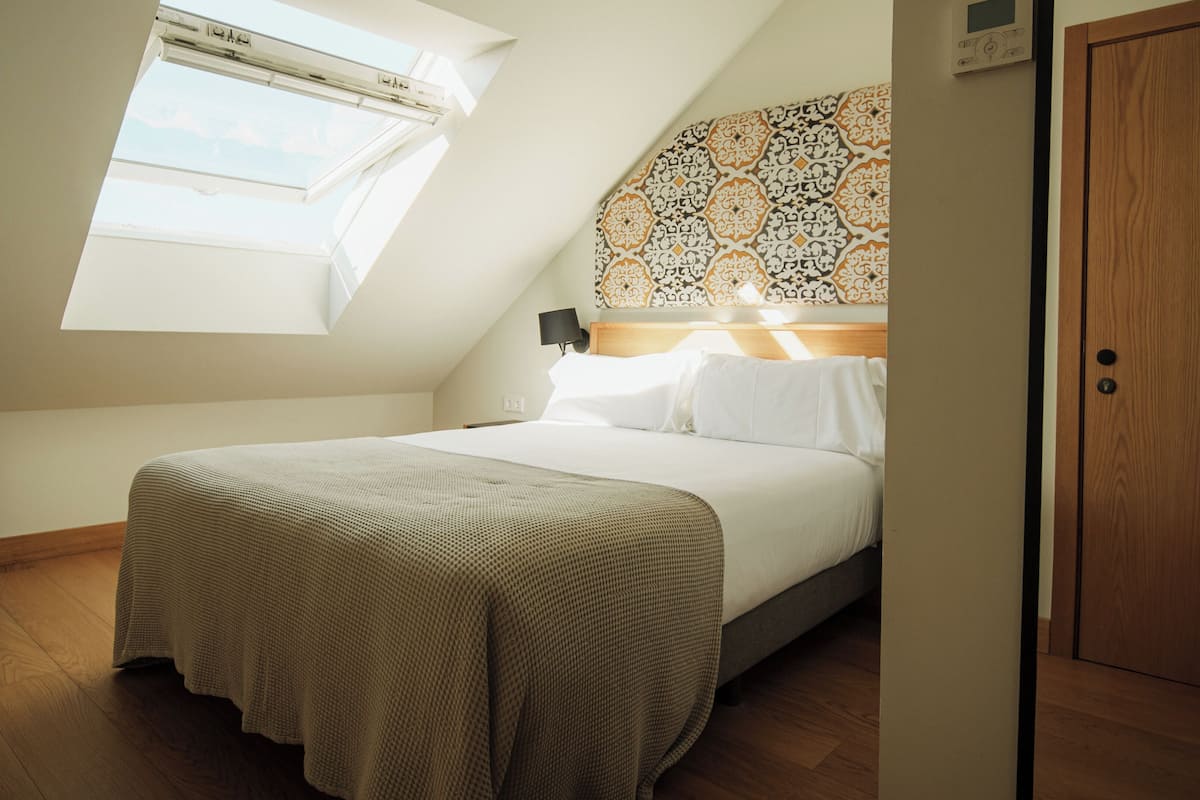 A bedroom with a sloped ceiling, a skylight, a bed with white linens, a patterned headboard, and a wooden door.
