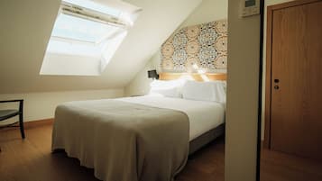 Quarto com teto inclinado, um claraboia, cama com roupa de cama branca, cabeceira estampada e porta de madeira.