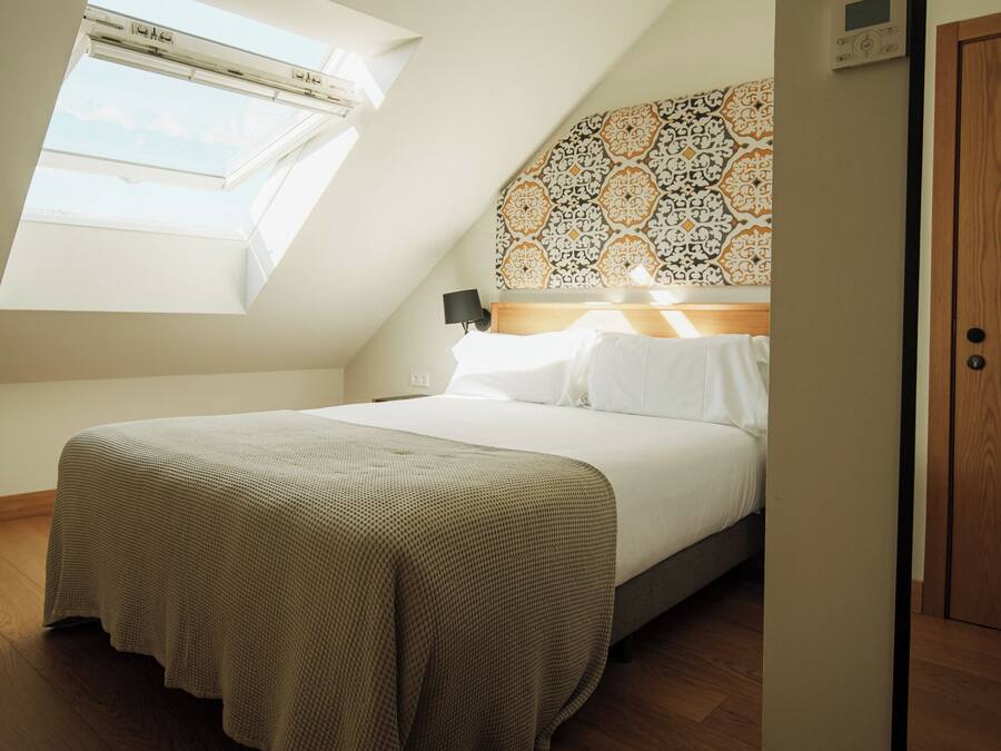 Quarto com teto inclinado, um claraboia, cama com roupa de cama branca, cabeceira estampada e porta de madeira.