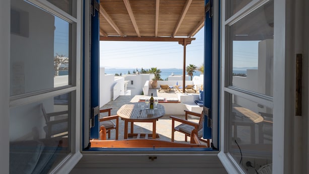 A balcony with a wooden table and chairs, a view of the sea, and white buildings in the background. - Beach House With Seaview (Naxos)