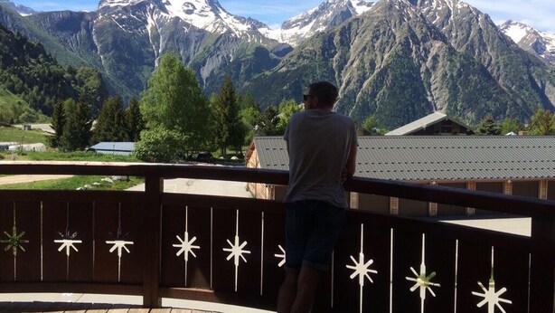 A person standing on a balcony overlooking a mountainous landscape with snow-capped peaks. - 2 Bed Apartment with great views backing onto the Diable ski lift (Les Deux Alpes)