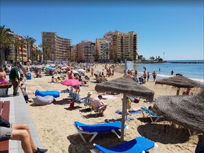 Guests can enjoy the beachside promenade with benches and lounge chairs, surrounded by beach umbrellas and a hotel complex in the background.