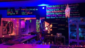 A bar with neon signs listing drinks and music, a counter with glasses, and a neon sign for 'X-Men Special'. - X-BAR and Bungalow (Trat)