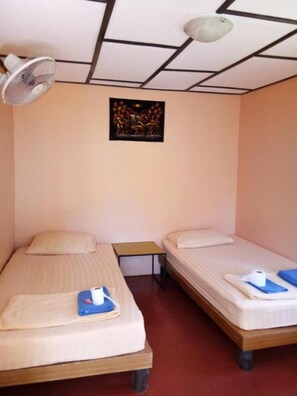 A small, single-room accommodation with two single beds, a small table, a ceiling fan, and a wall-mounted picture. - X-BAR and Bungalow (Trat)