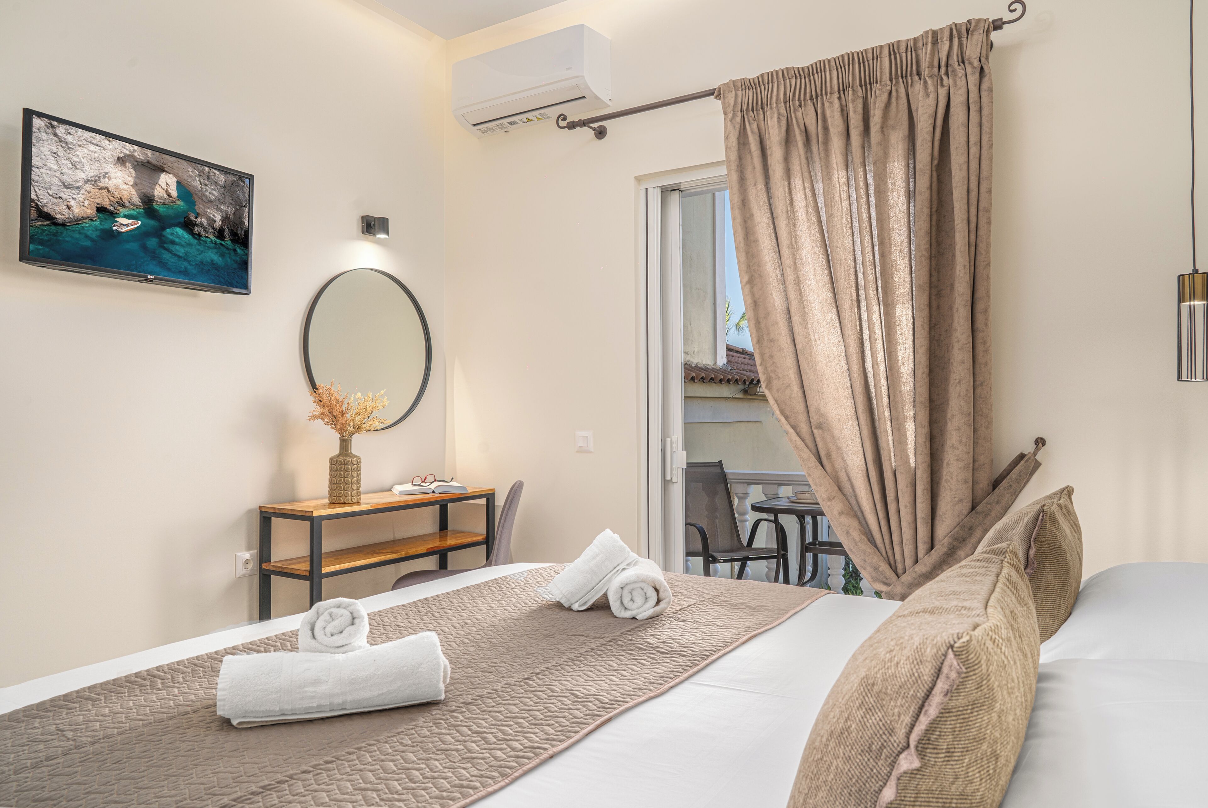 Habitación de hotel con cama, escritorio y silla, televisor, ventana con cortinas y vistas al balcón.