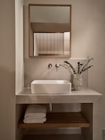 A bathroom with a rectangular sink, a mirror, and a wooden shelf with towels.
