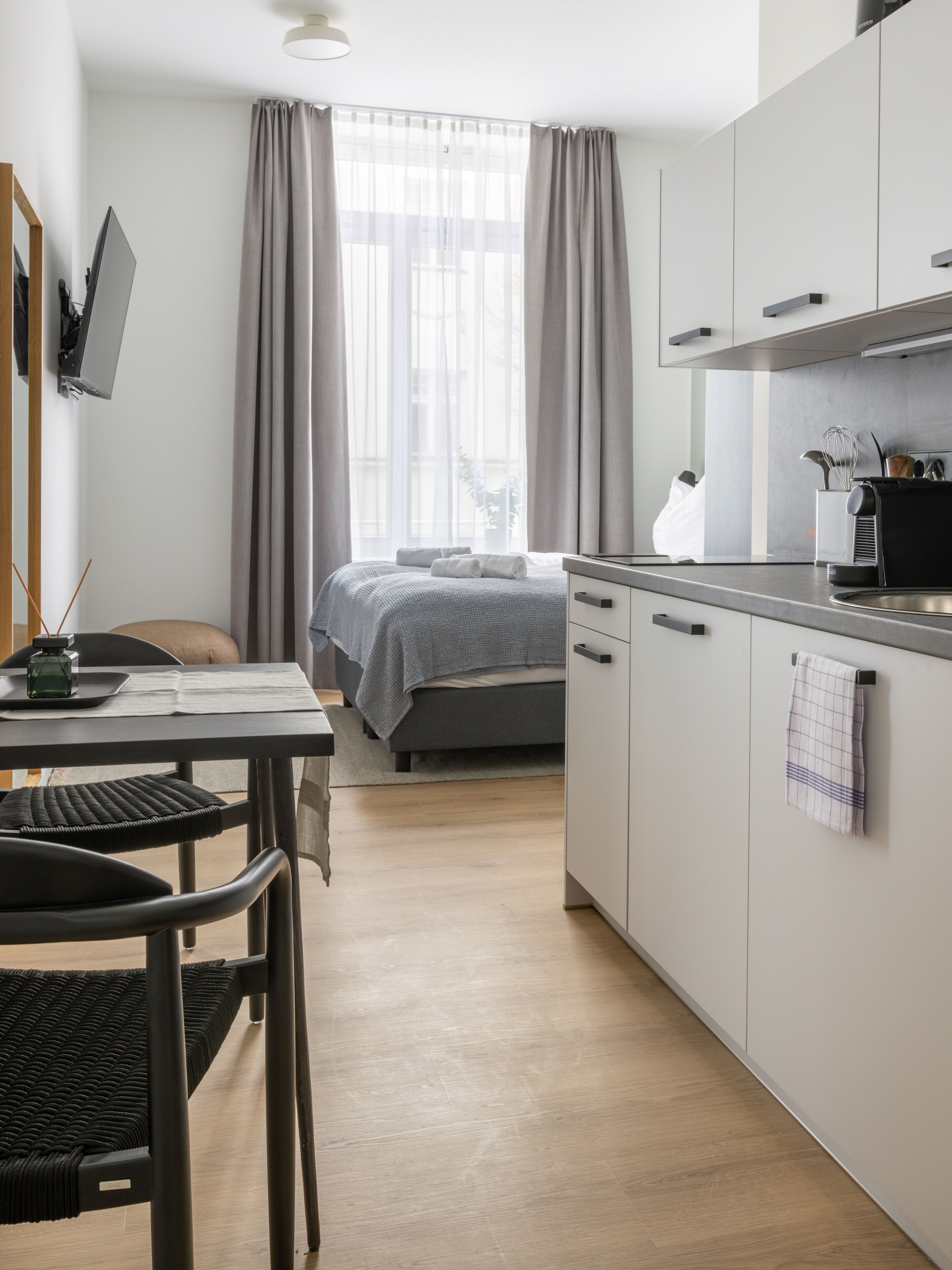 A modern hotel room with a bed, a desk, a chair, a kitchenette, and a large window with curtains.