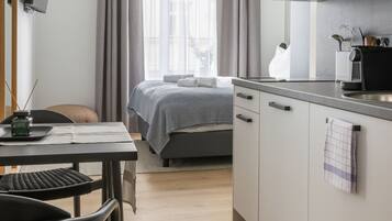 A modern hotel room with a bed, a desk, a chair, a kitchenette, and a large window with curtains.