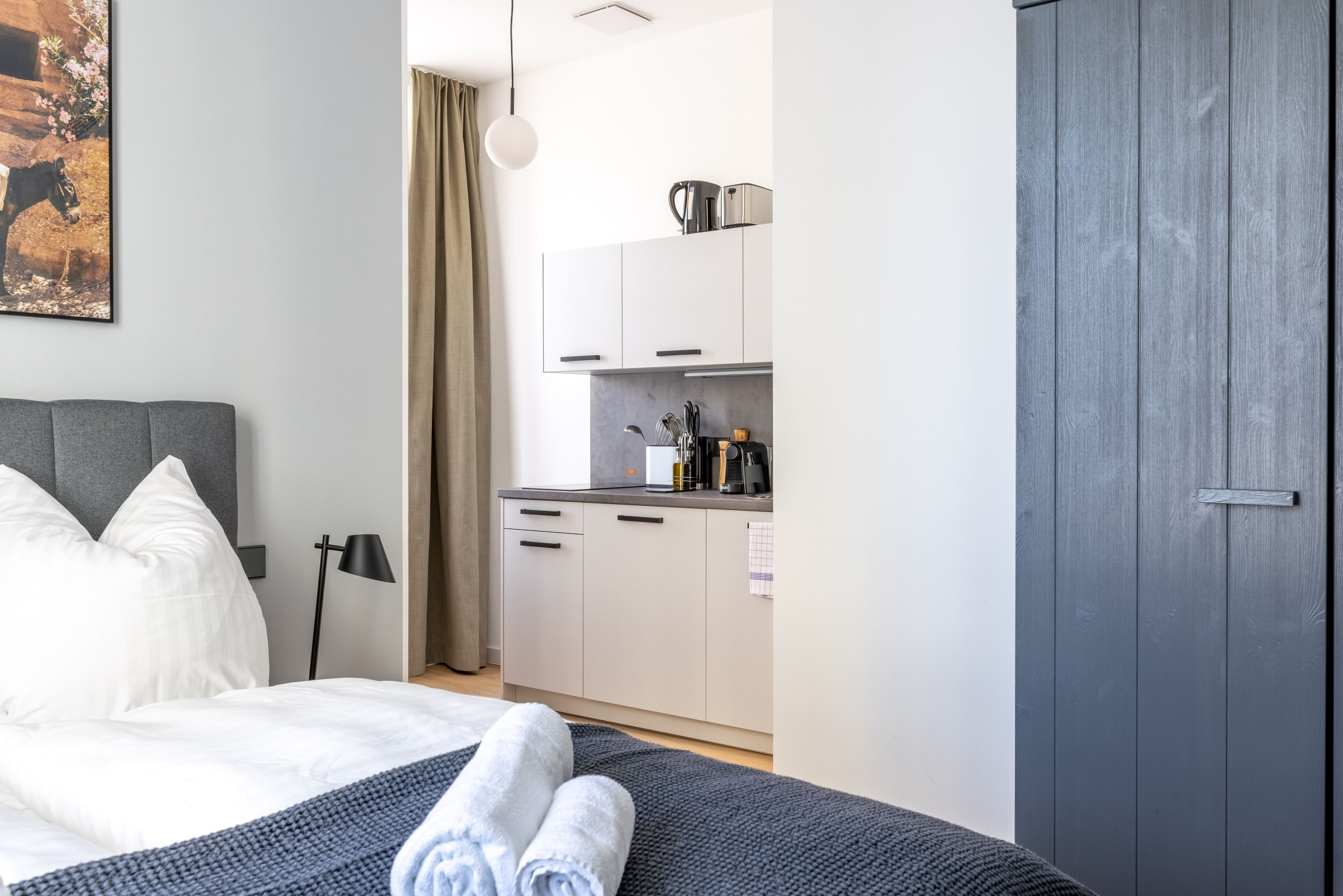 A modern hotel room with a neatly made bed, a small kitchenette, and a grey wardrobe.