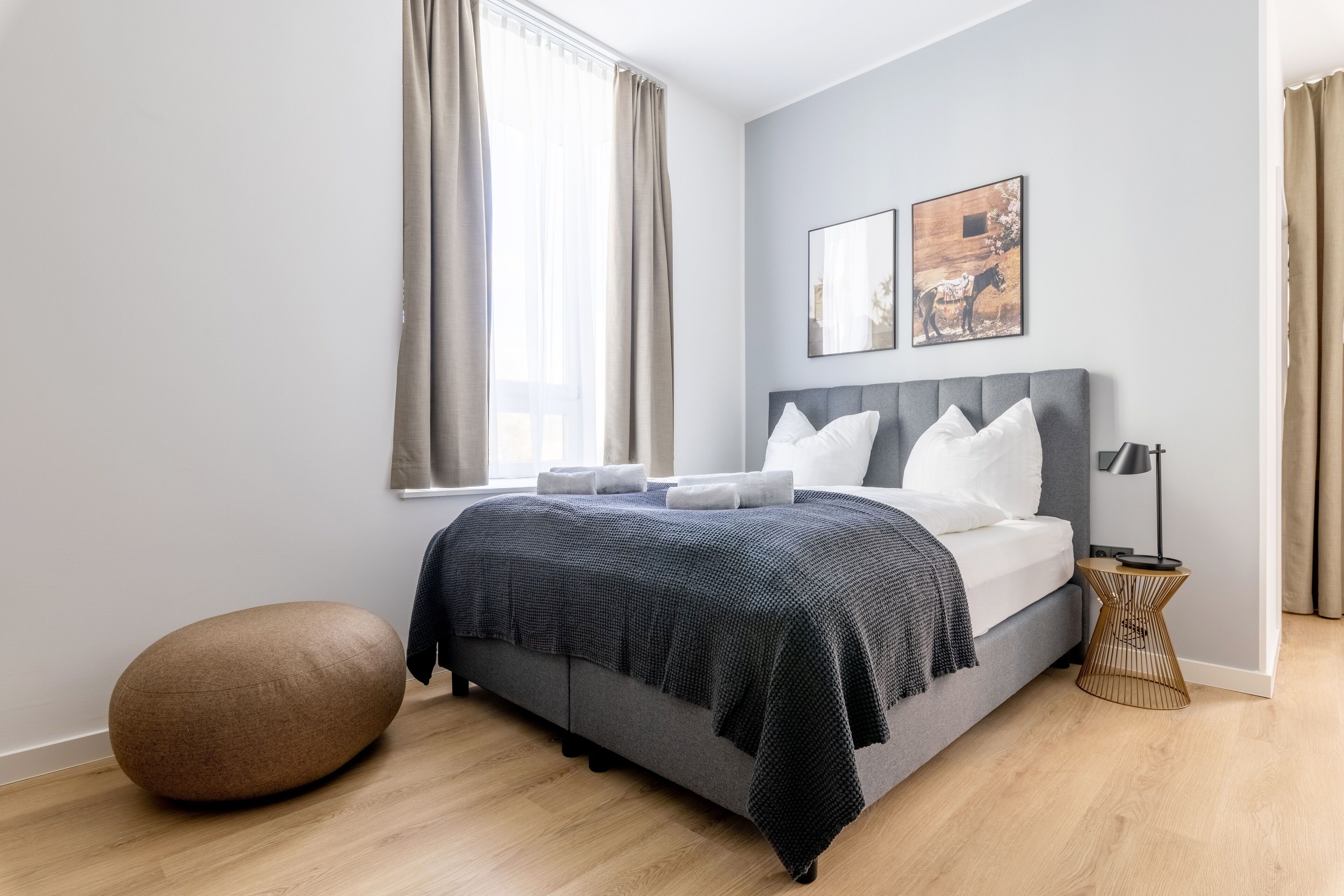 A modern bedroom with a bed, a round ottoman, and two wall-mounted artworks.