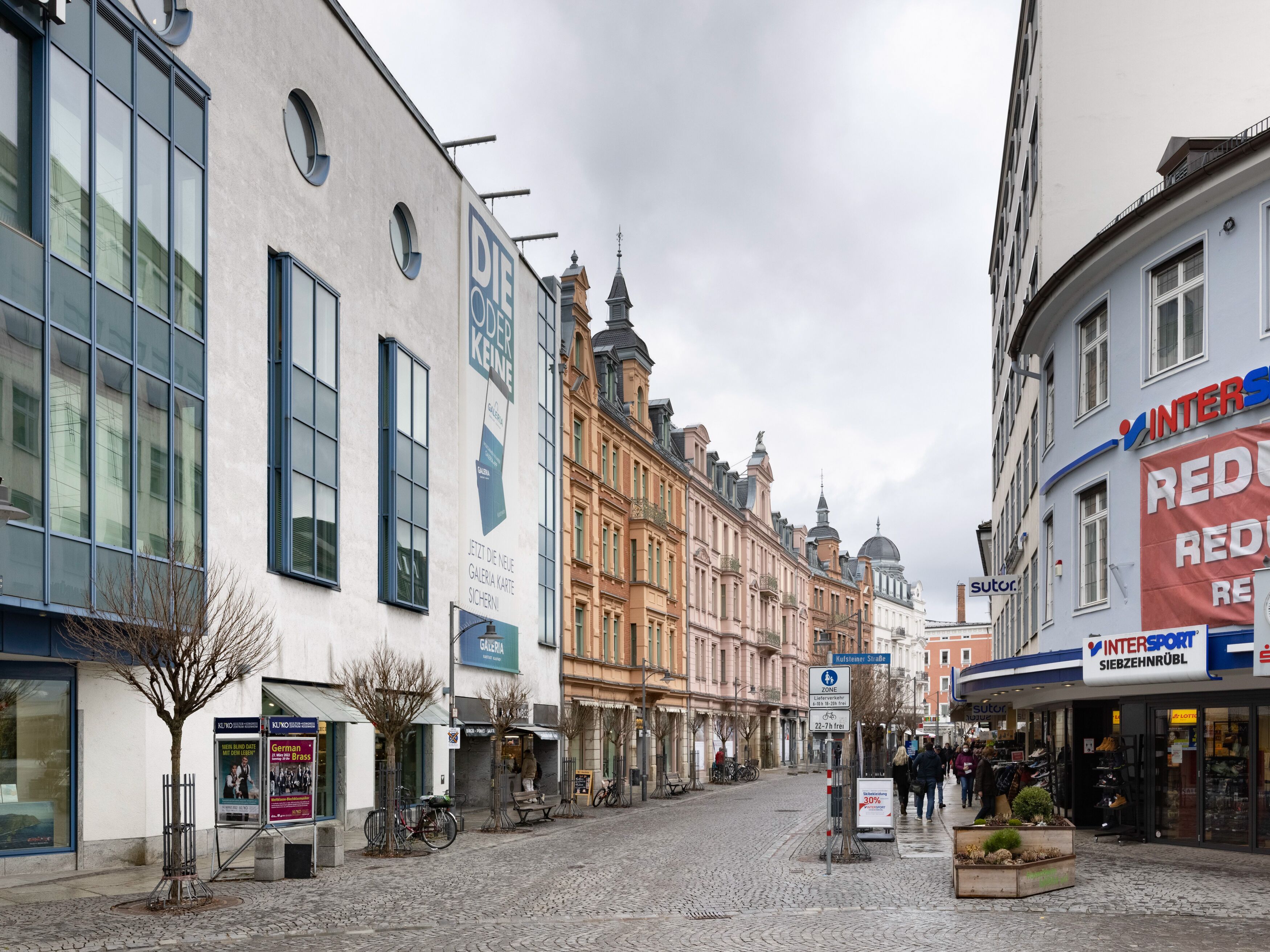 Una calle adoquinada flanqueada por edificios históricos, incluyendo un moderno edificio de cristal y una construcción tradicional con un letrero que dice 'INTERSPORT SIEBZEHNRÜBL'.