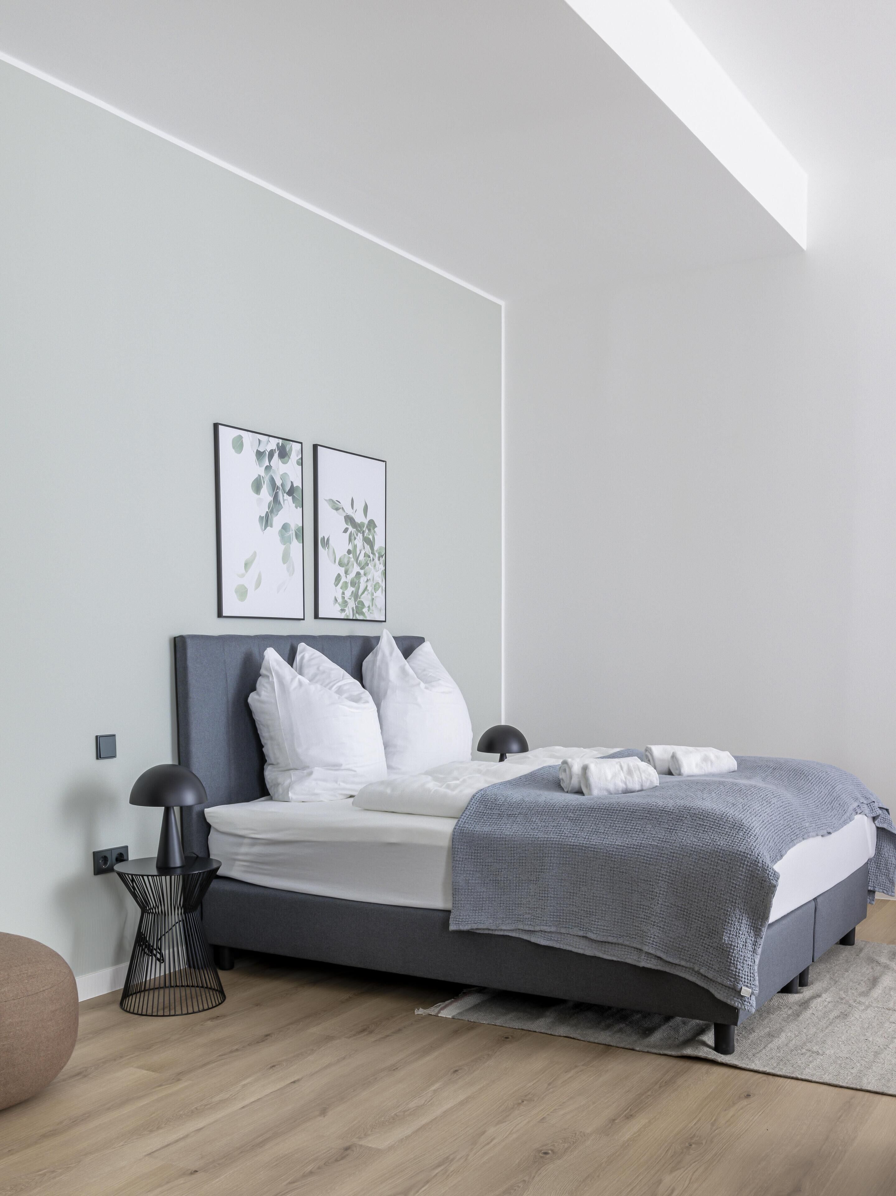 A modern bedroom with a grey bed, white pillows, a grey throw blanket, and two framed botanical prints on the wall.