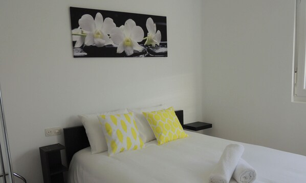 Quarto de hotel com cama, almofadas, pintura de orquídeas e ícones indicando comodidades como estacionamento gratuito, localização, Wi-Fi, controlo de desinfeção O3 e conta Netflix gratuita.