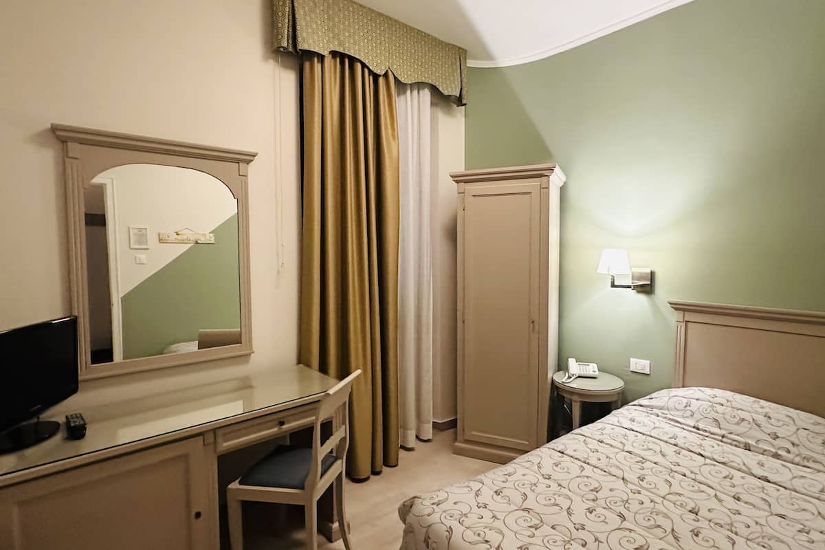 A hotel room with a bed, a desk with a chair, a mirror, a wardrobe, a bedside table with a lamp, and a telephone.