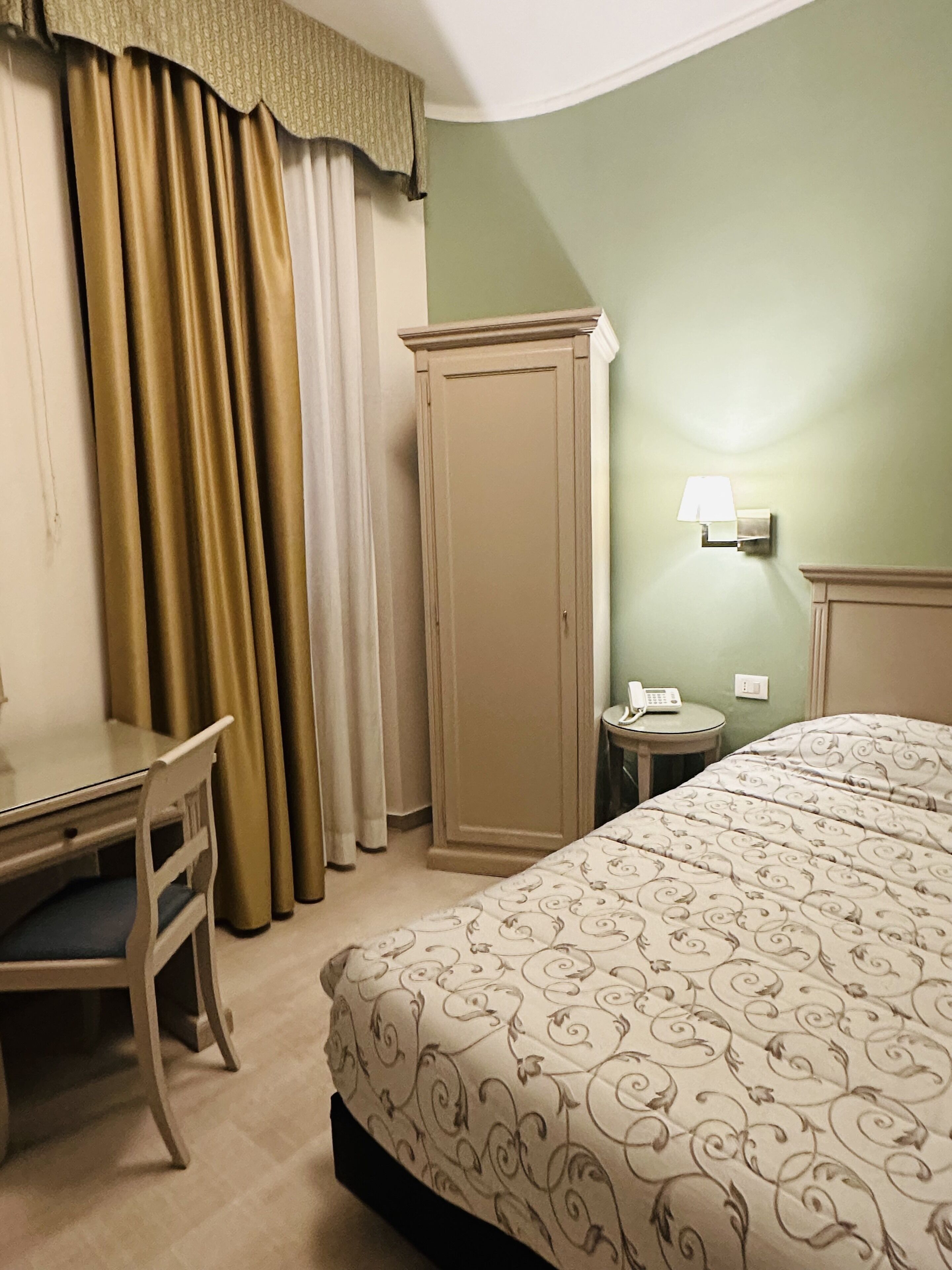 A hotel room with a bed, a desk, a chair, a wardrobe, and a bedside table with a lamp.