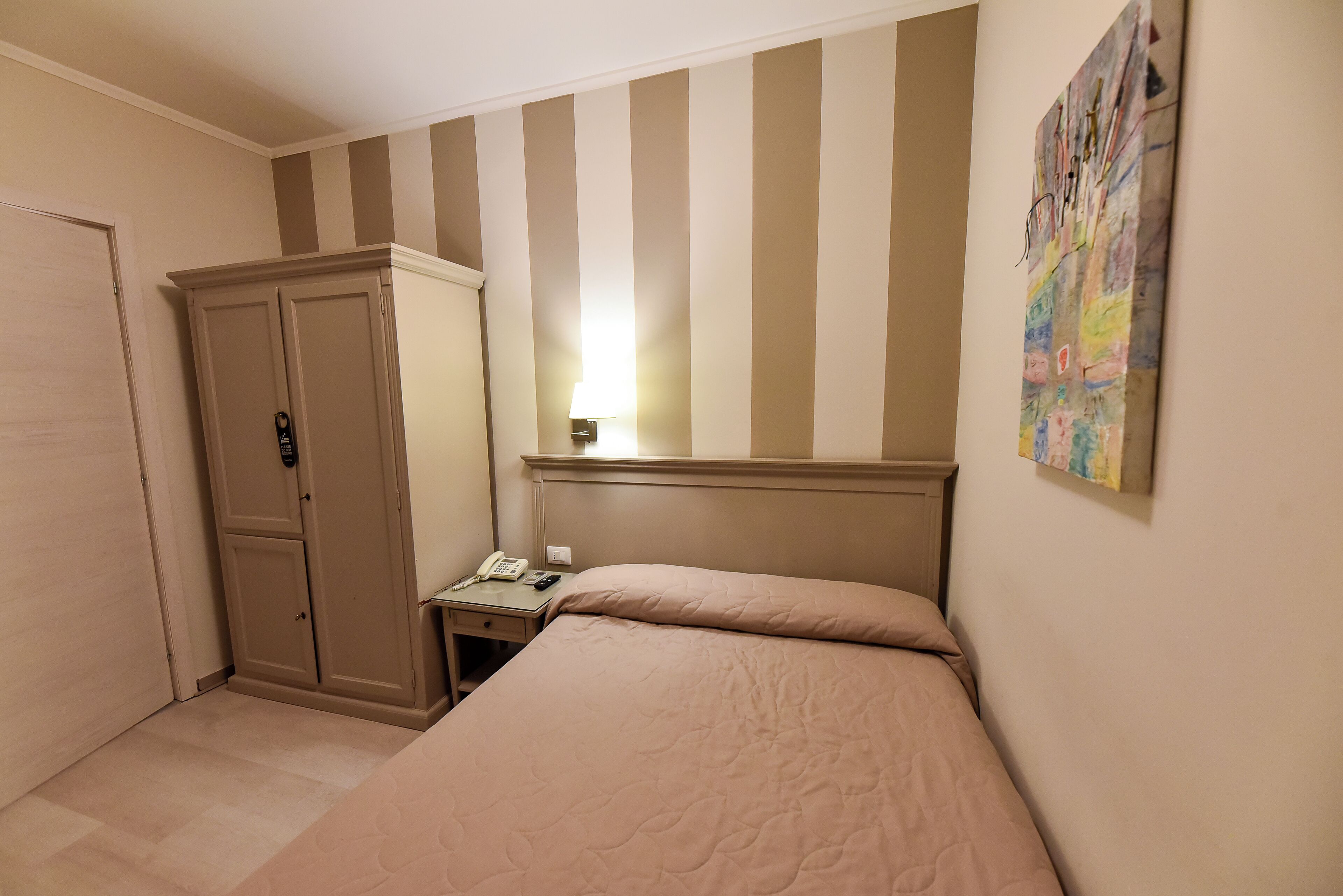 A hotel room with a bed, a wardrobe, a desk with a lamp, and a wall-mounted artwork.