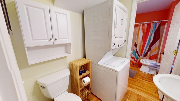 Un baño pequeño y funcional con inodoro, un estante de madera con artículos de aseo, un lavabo blanco y cortina de ducha.
