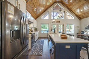 A spacious kitchen with a large island, wooden ceiling, and large windows offering views of the outdoors.