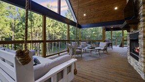 A covered outdoor deck with a stone fireplace, wooden furniture, and a view of trees. - A Rustic Cabin with a Modern Twist w Hot Tub,Fire Pit,Fireplace,Near Lake Sleep7 (Broken Bow)