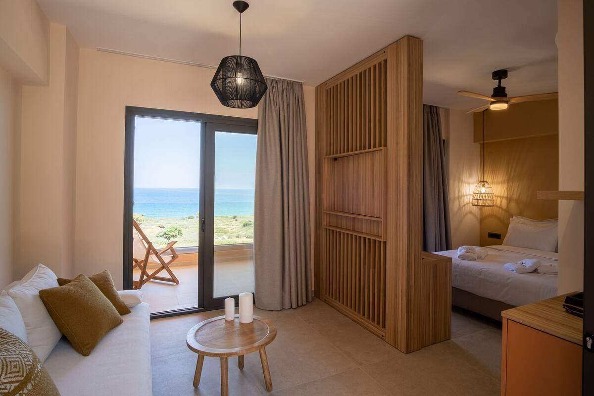 A modern hotel room with a large sliding glass door leading to a balcony with a view of the sea. There is a bed with white linens, a wooden sideboard, and a round wooden table with two candles.