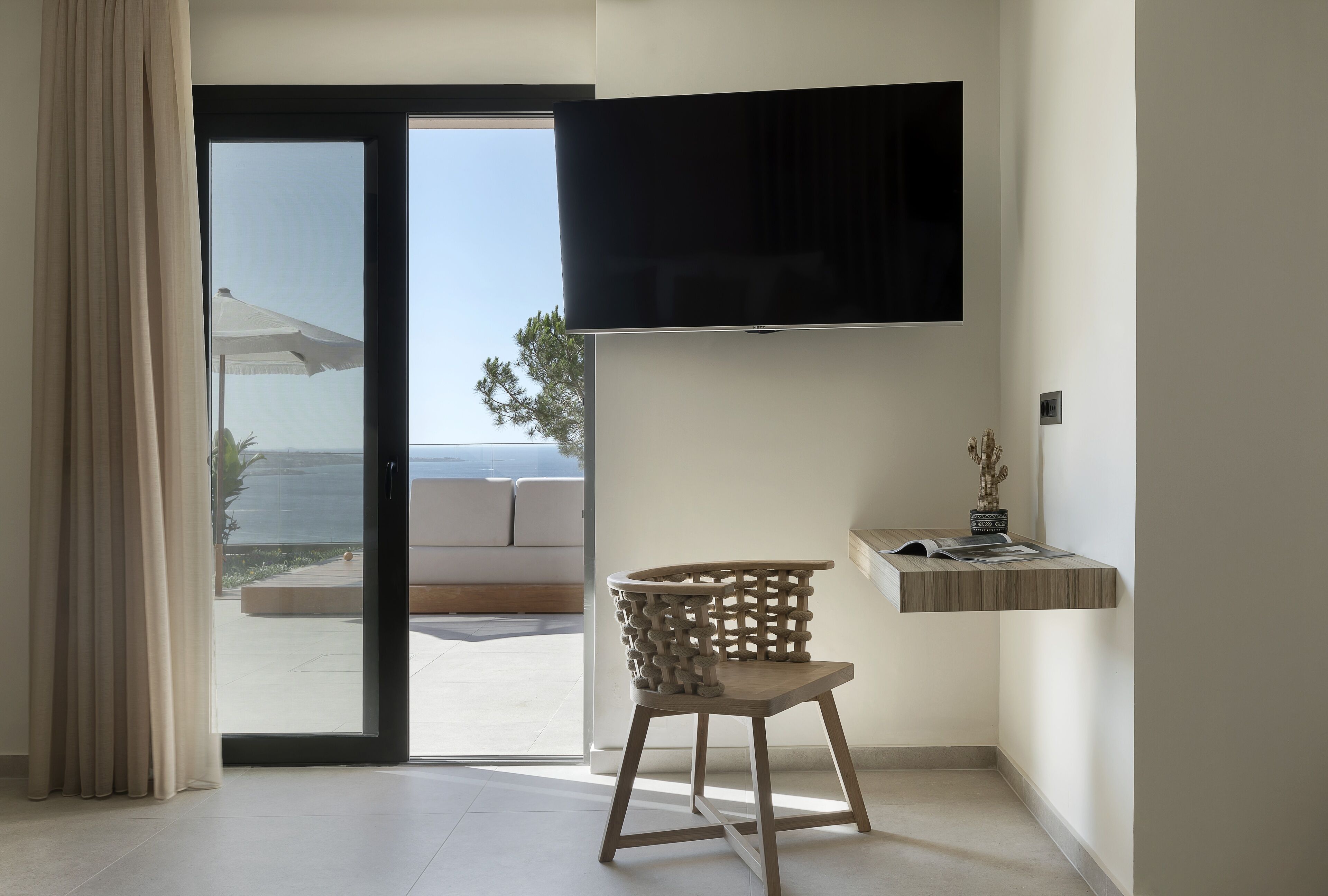 Une chambre moderne dotée d’une porte-fenêtre coulissante donnant sur un balcon, d’un téléviseur à écran plat fixé au mur, d’une chaise en bois et d’une petite étagère en bois ornée d’objets décoratifs.