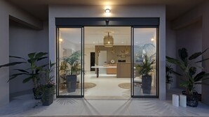 A modern hotel entrance with a glass door, potted plants, a reception desk, and a hanging light fixture.