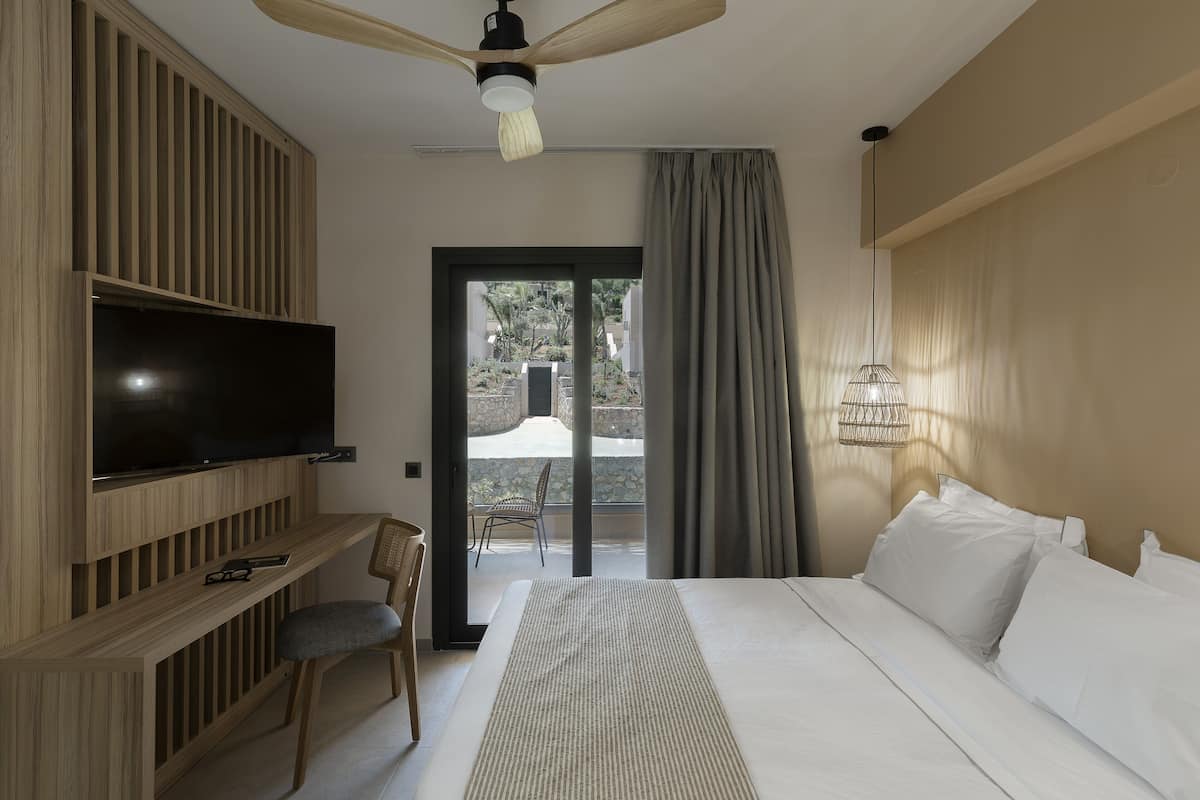 A modern hotel room with a large bed, a flat-screen TV mounted on a wooden panel, a ceiling fan, and a view of the outdoor area through a sliding door.