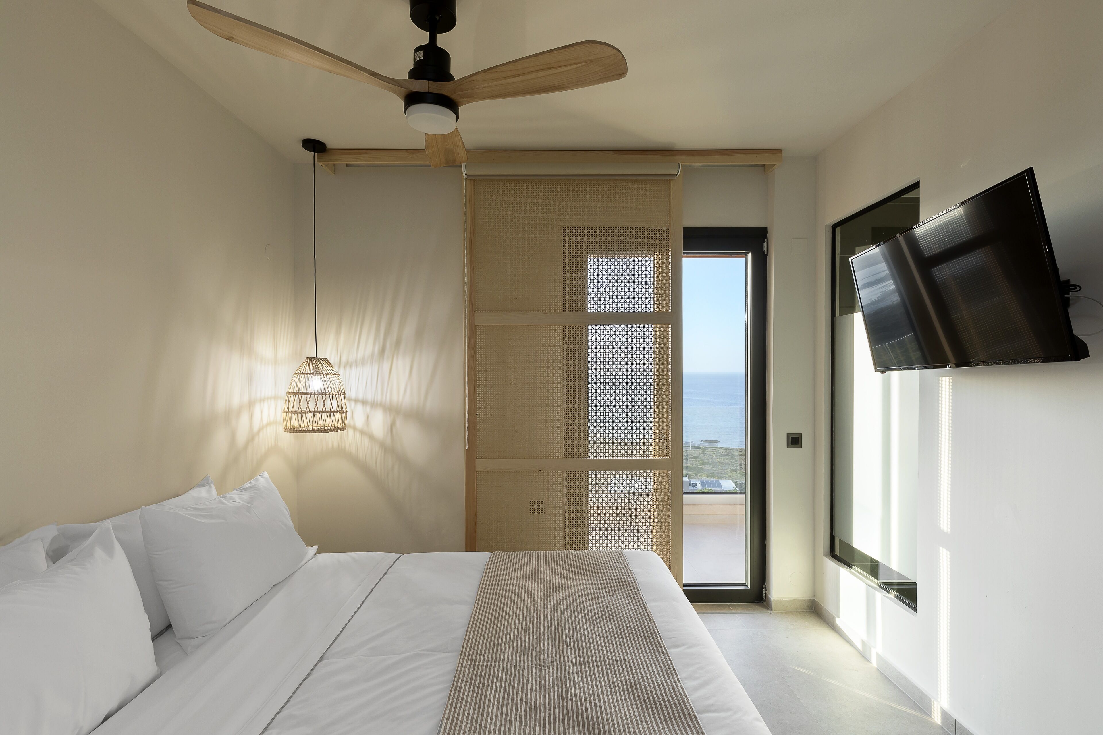 A bedroom with a bed, a ceiling fan, a TV mounted on the wall, and a view of the sea through a window.