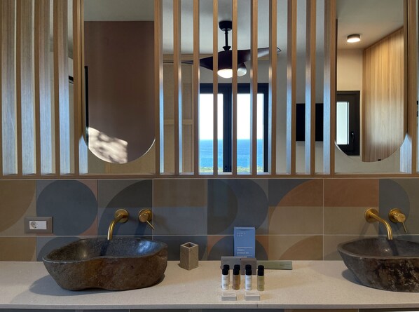 A bathroom with two stone sinks, a ceiling fan, and a view of the ocean through large windows. - The Nest Resort (Hersonissos)