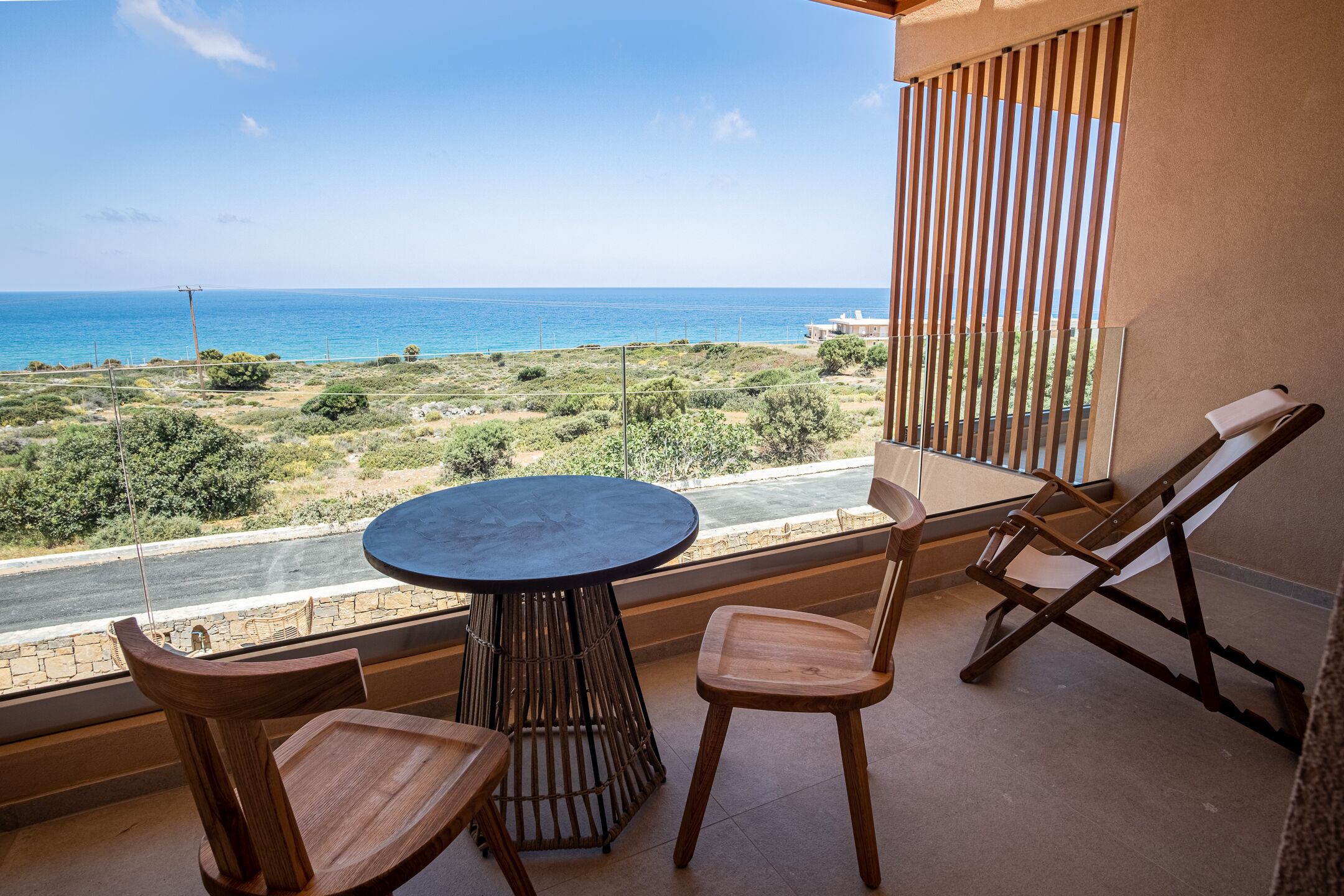 A balcony with a round table and two wooden chairs, overlooking a coastal landscape with a clear view of the sea.