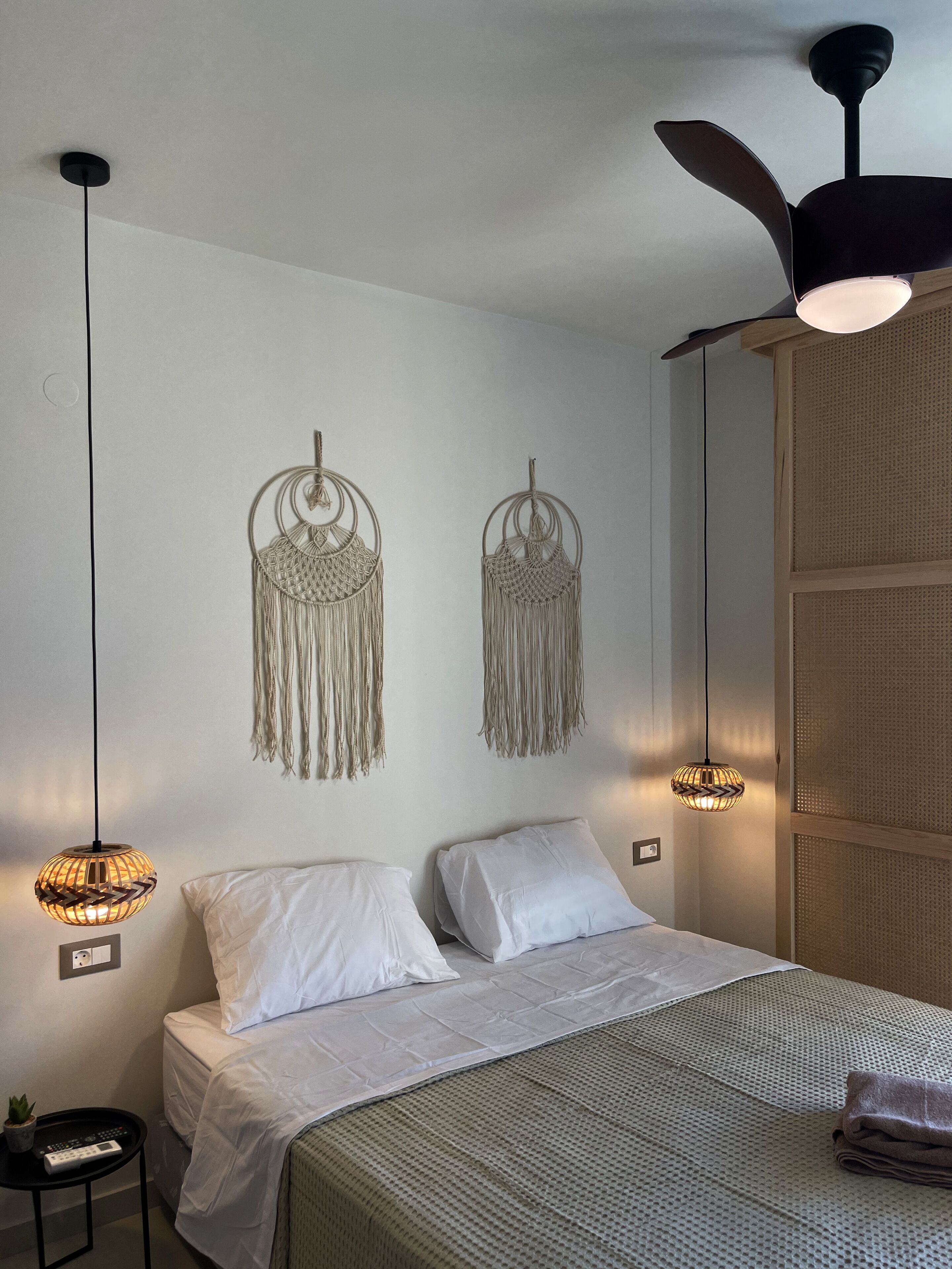 A bedroom with a bed, two hanging macrame wall decorations, a ceiling fan, and two wall-mounted lamps.