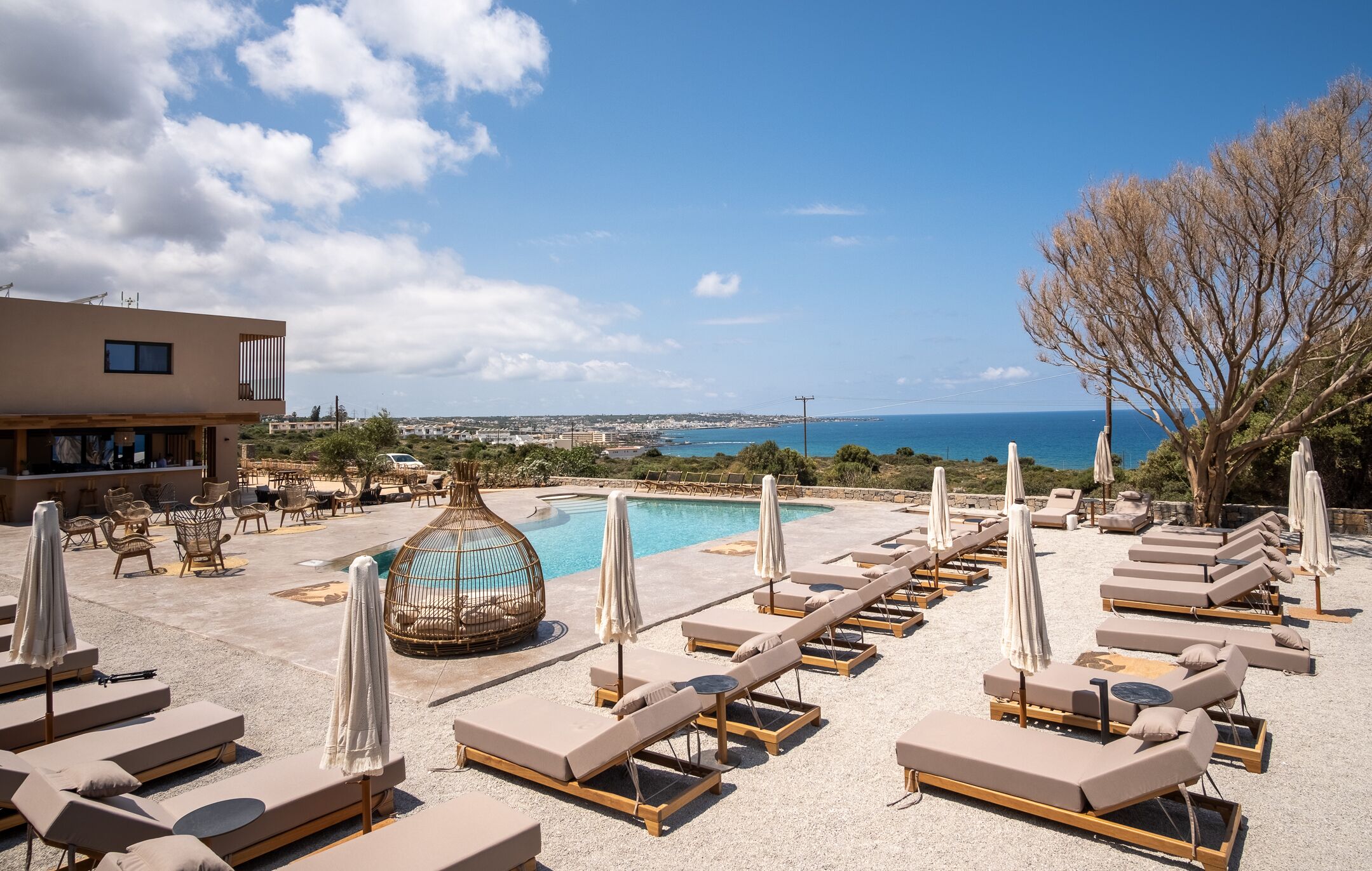 A rooftop pool area with lounge chairs and umbrellas, a large decorative vase, and a view of the sea and distant buildings.