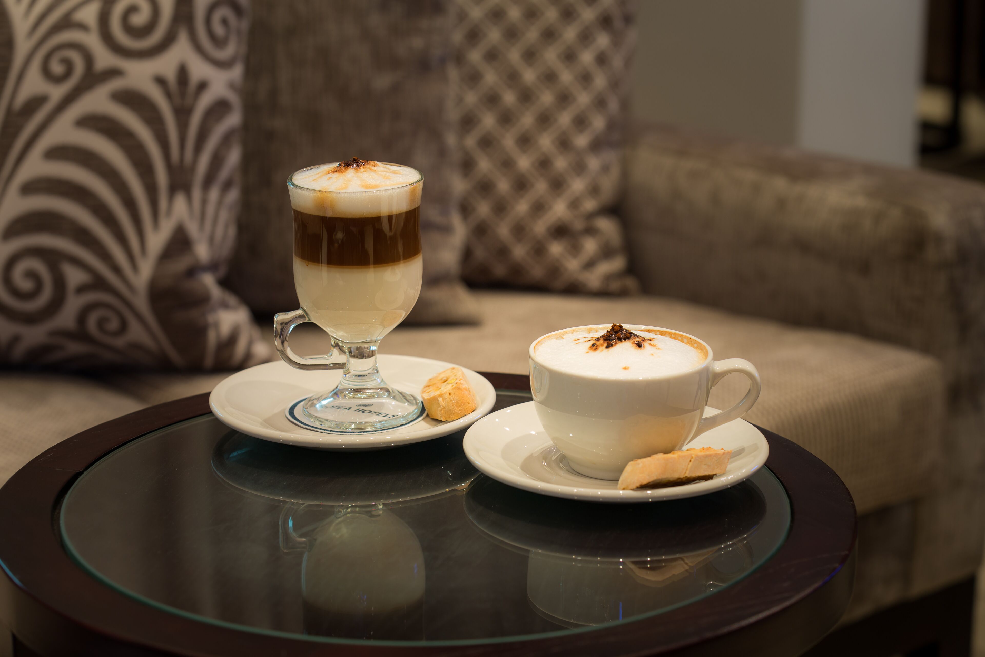 Two cups of coffee on a glass table with a sofa in the background.