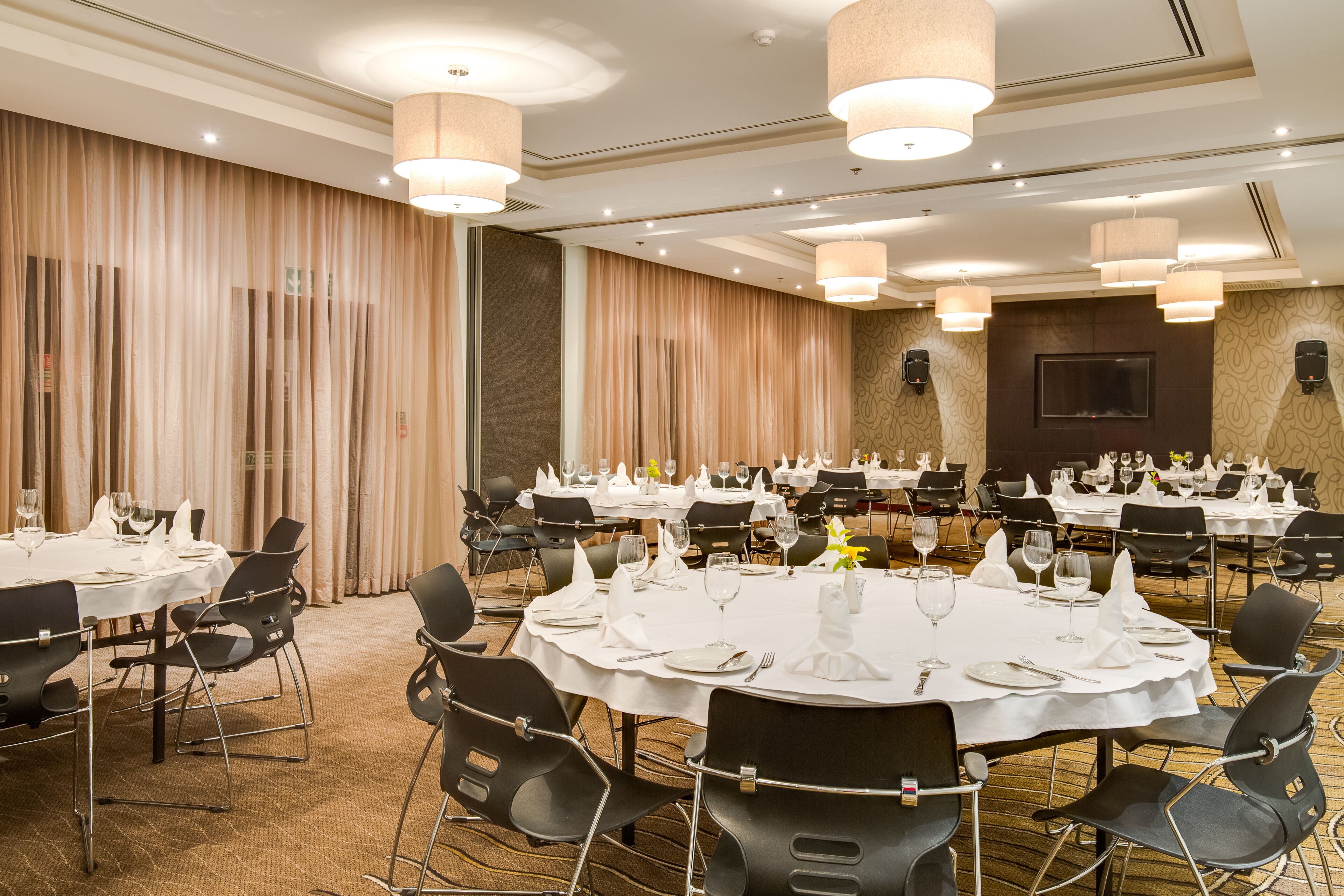A banquet hall with round tables set for dining, black chairs, and hanging pendant lights.