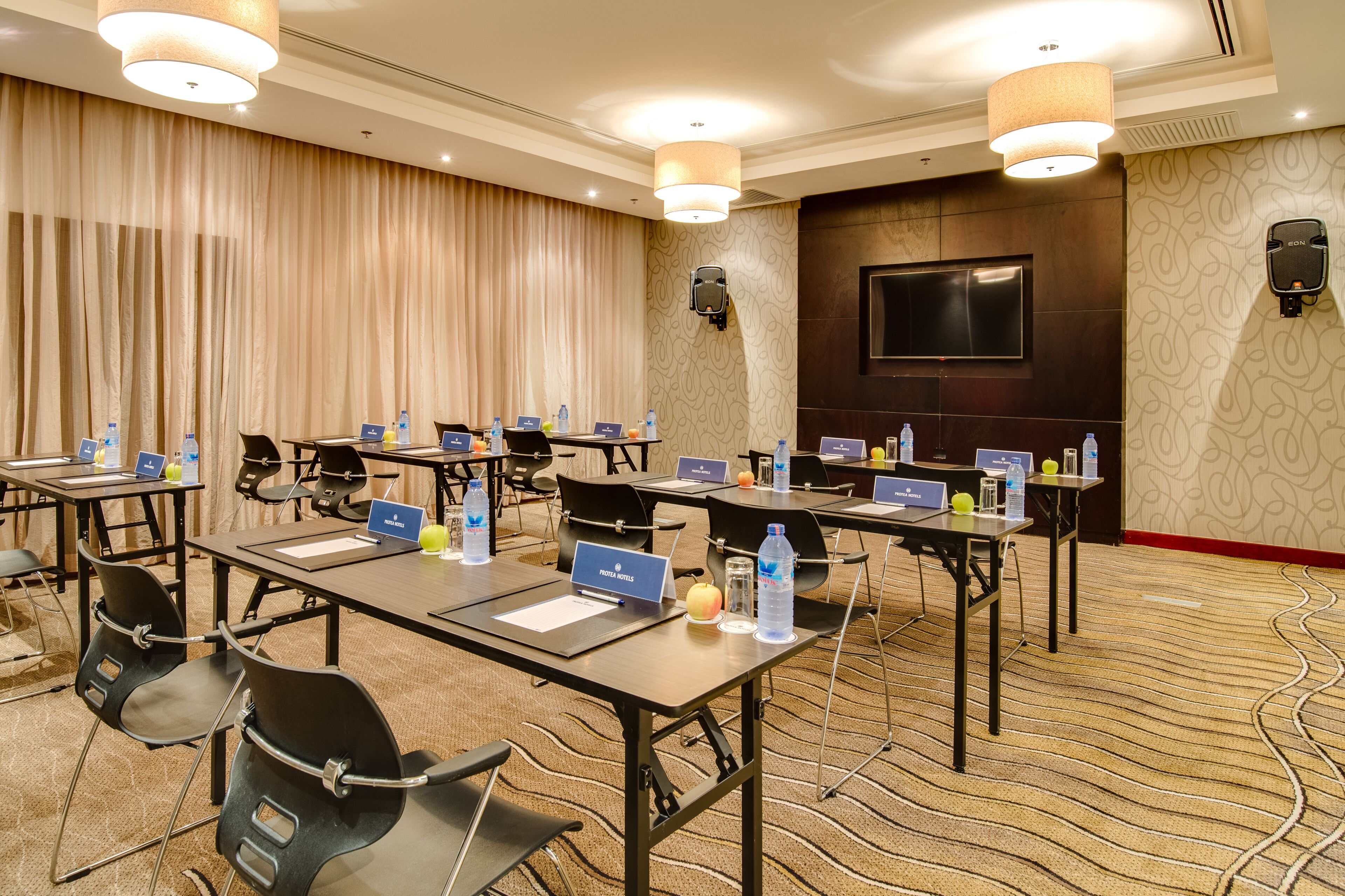 Un salón de conferencias con varias mesas, sillas, tarjetas de identificación, computadoras portátiles, botellas de agua y un televisor en la pared.