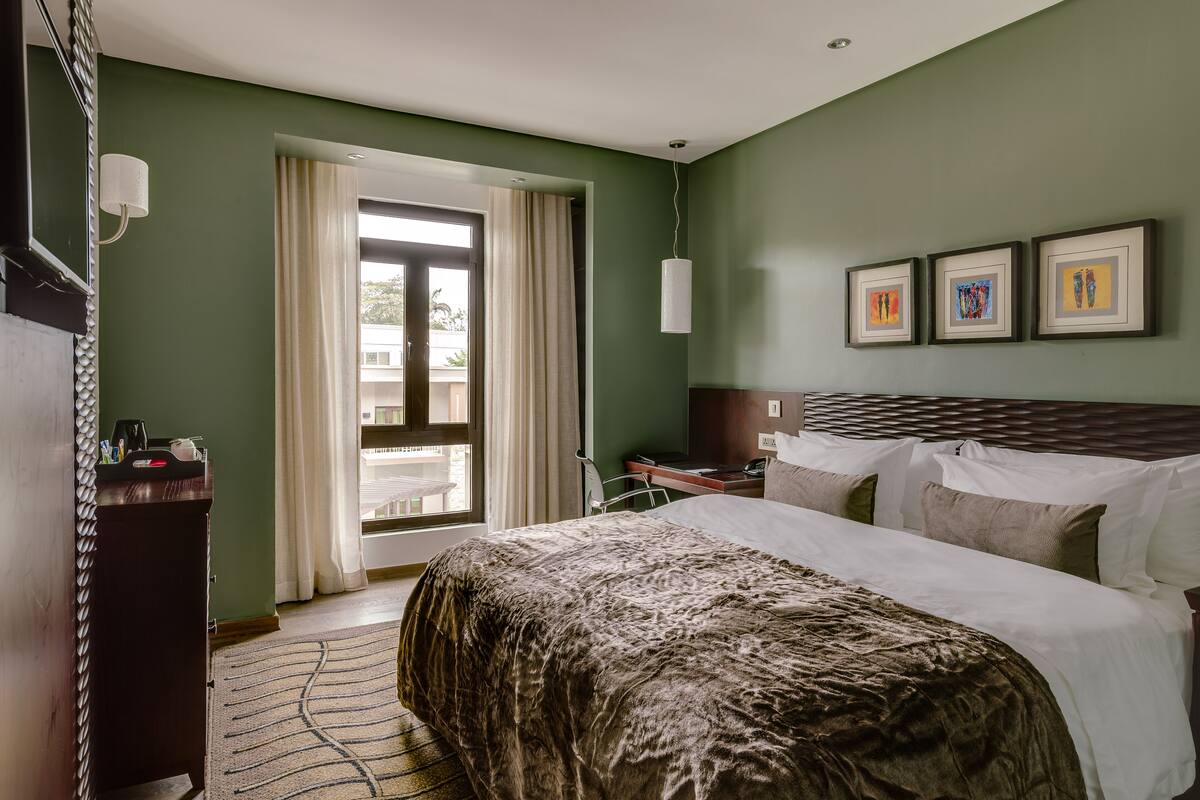 Una habitación de hotel con una cama grande, un escritorio, una silla, una ventana con cortinas y obras de arte en la pared.